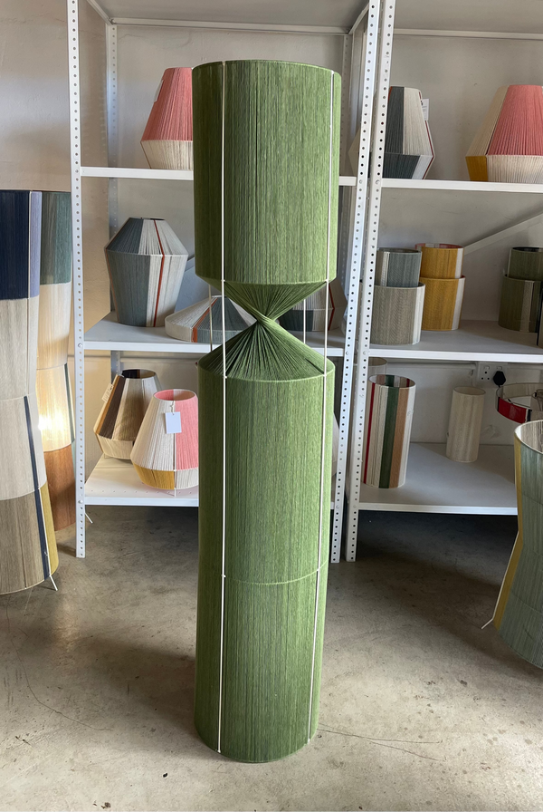 Green cylindrical lamp with patterned design on a concrete floor with shelves of various lamps in the background.